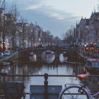 Evening view of an Amsterdam canal with a bridge and city lights reflecting in the water, framed by bicycles in the foreground