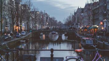 Evening view of an Amsterdam canal with a bridge and city lights reflecting in the water, framed by bicycles in the foreground