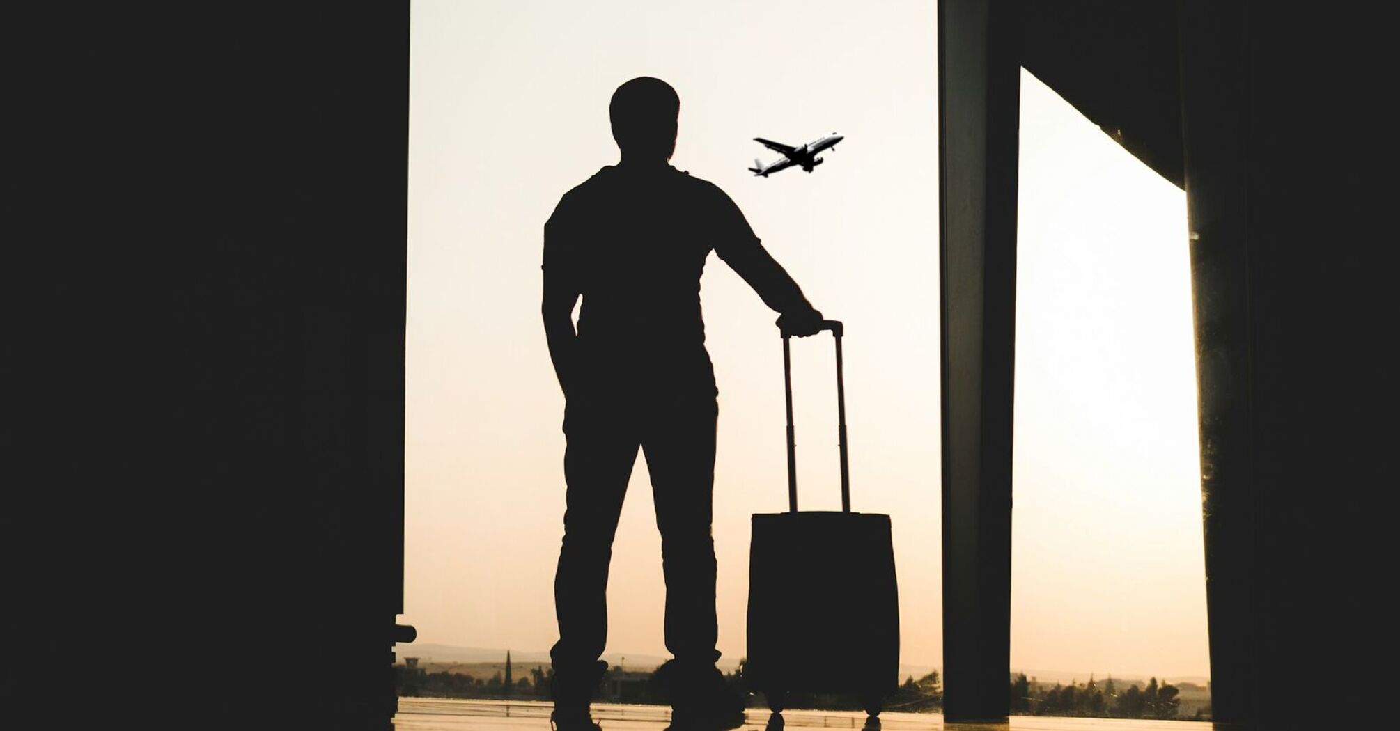 Silhouette of a traveler with a suitcase watching a plane take off at the airport