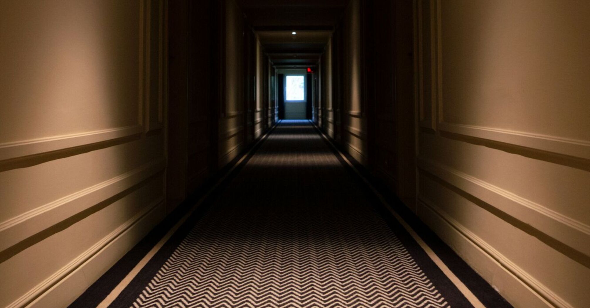 A dimly lit hotel hallway with a patterned carpet and doors on each side, leading to a glowing exit at the far end