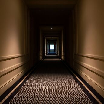 A dimly lit hotel hallway with a patterned carpet and doors on each side, leading to a glowing exit at the far end