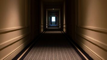 A dimly lit hotel hallway with a patterned carpet and doors on each side, leading to a glowing exit at the far end
