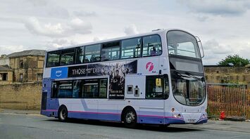 Double-decker First bus with an advertisement on the side, parked near an old building under a cloudy sky