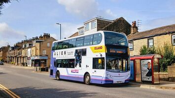 A double-decker First Bus parked at a bus stop in a residential area, with clear signage and advertisements on its side