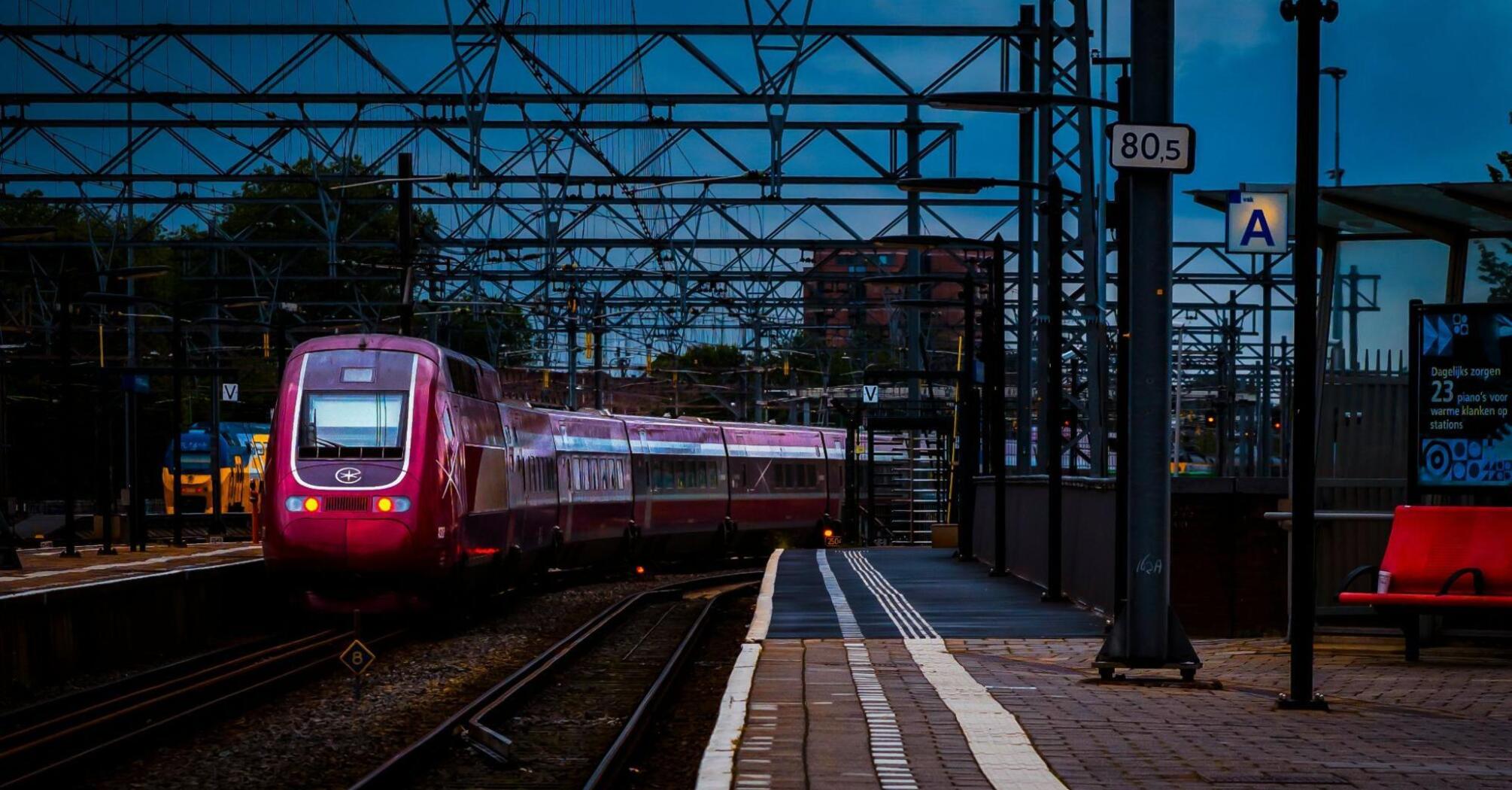 A high-speed red train on a dimly lit station platform, surrounded by overhead wires and structures, preparing for departure