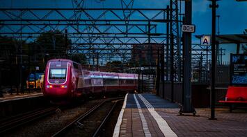 A high-speed red train on a dimly lit station platform, surrounded by overhead wires and structures, preparing for departure