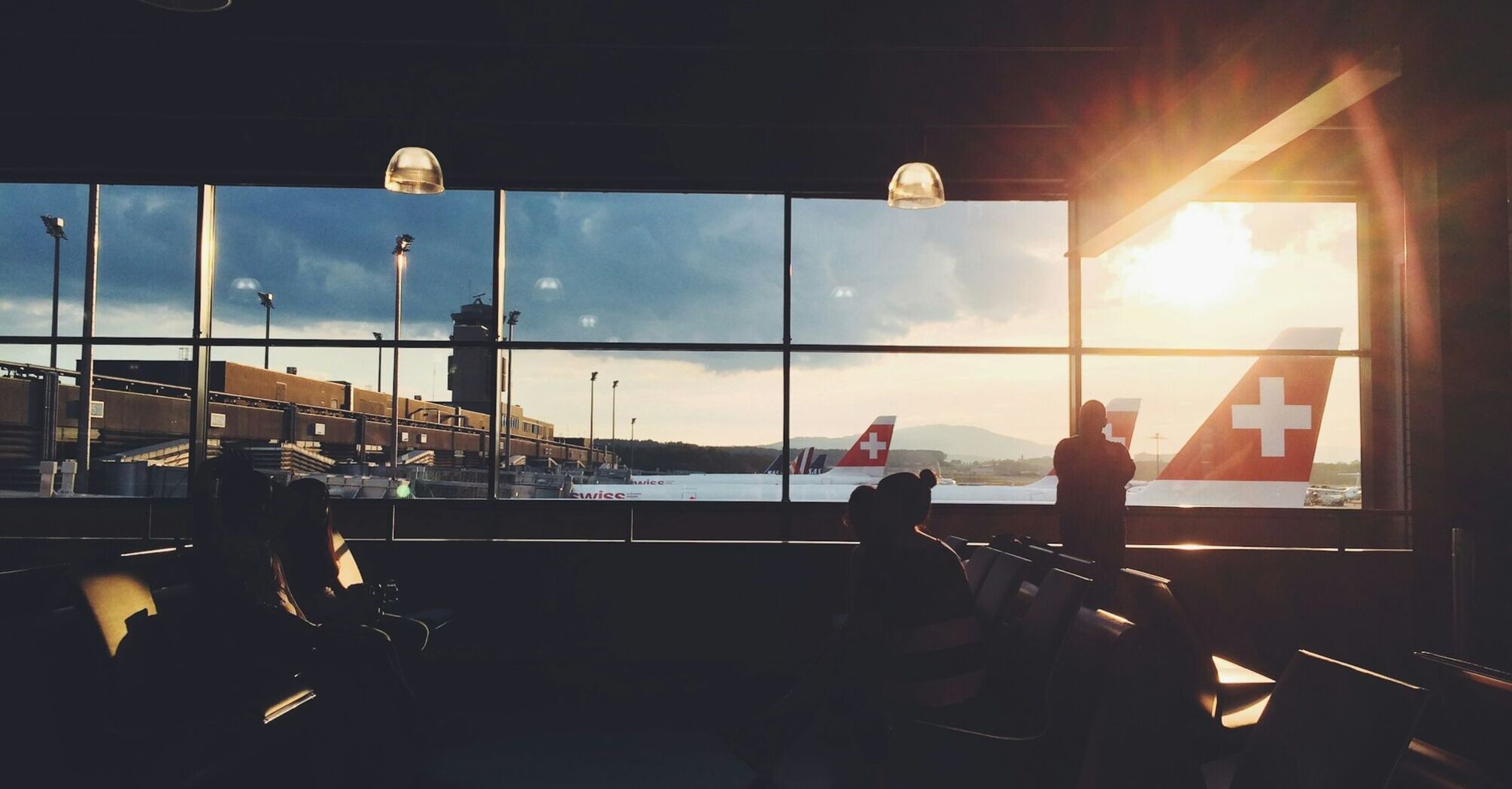 Passengers at an airport terminal with SWISS planes visible through large windows during sunset