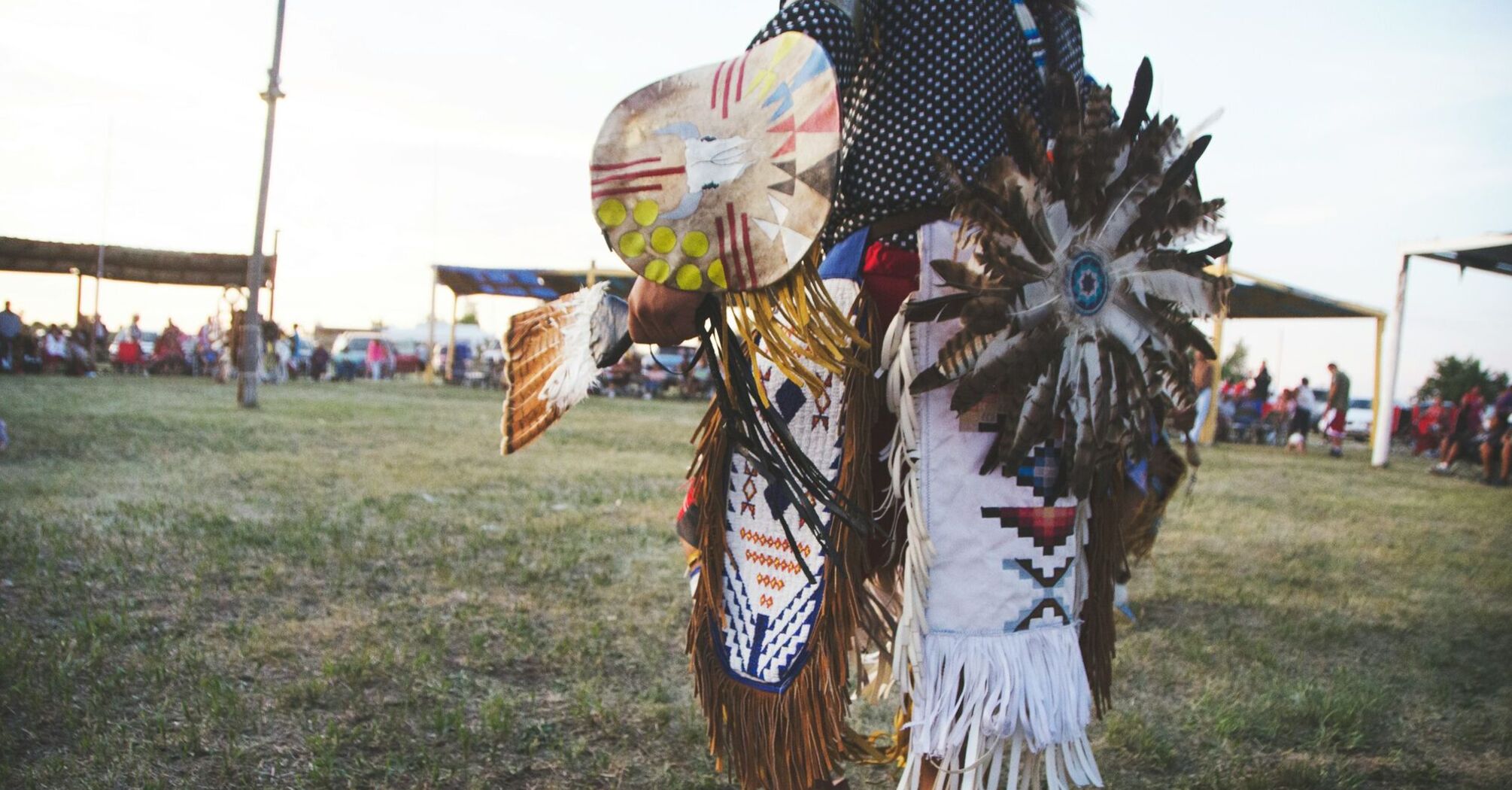 A dancer in traditional Indigenous attire with feathered accessories and painted shield performs during a cultural event