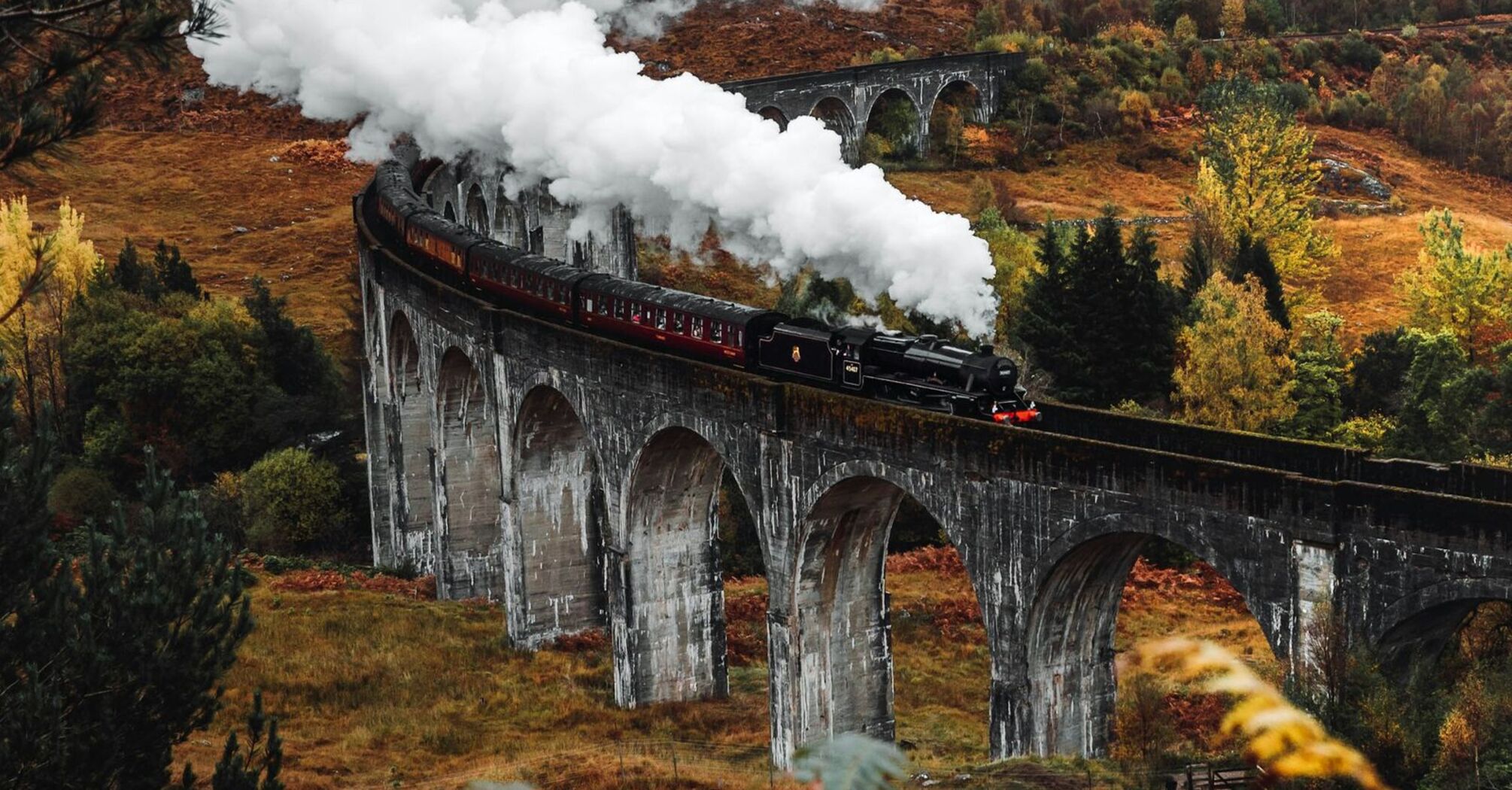 A historic steam train crosses a scenic viaduct surrounded by autumn foliage, emitting white smoke against a backdrop of hills