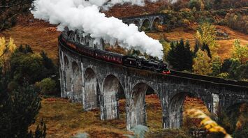 A historic steam train crosses a scenic viaduct surrounded by autumn foliage, emitting white smoke against a backdrop of hills