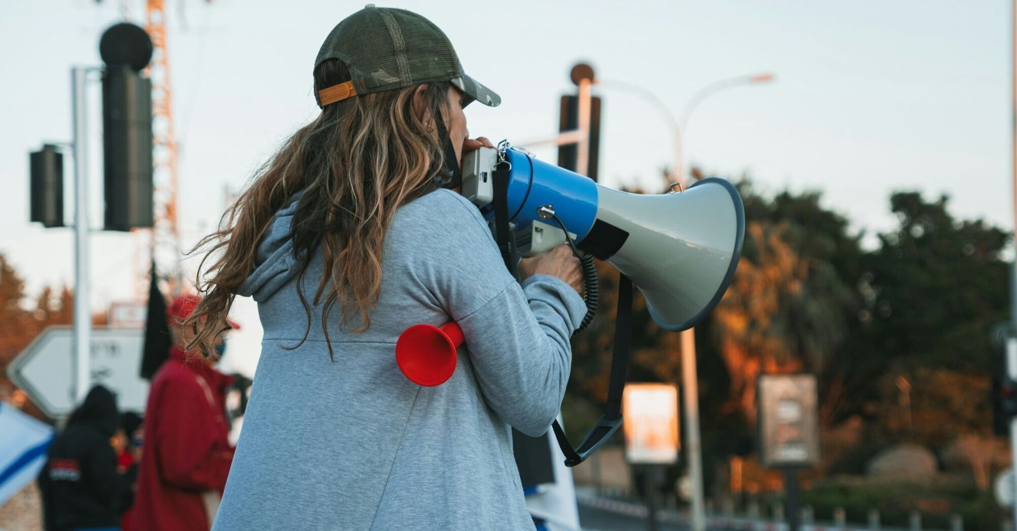 A person with long hair wearing a cap speaks into a megaphone at an outdoor gathering, highlighting a protest or rally scene