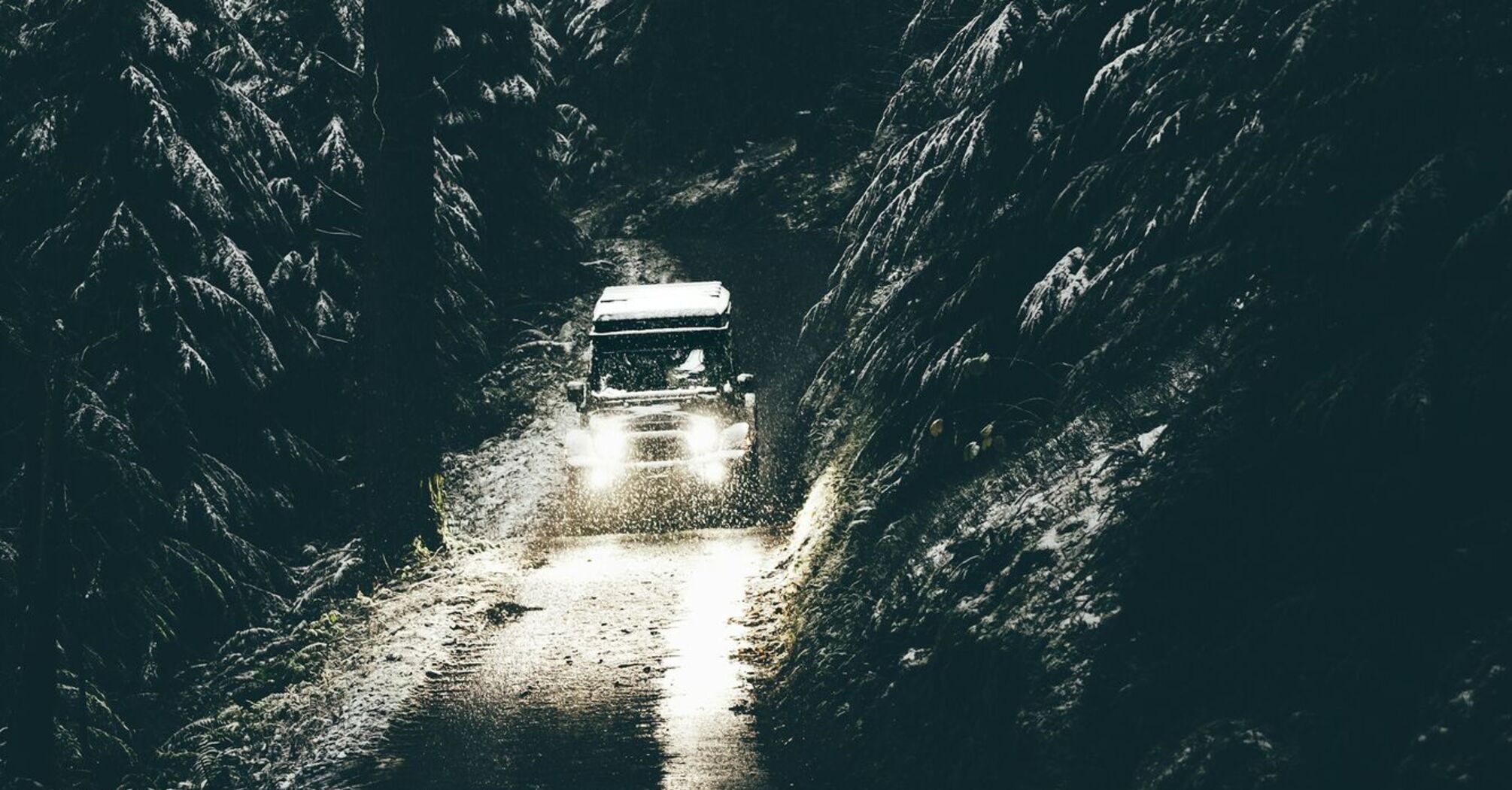 A car drives through a narrow, snowy forest road with dim lighting in Wales