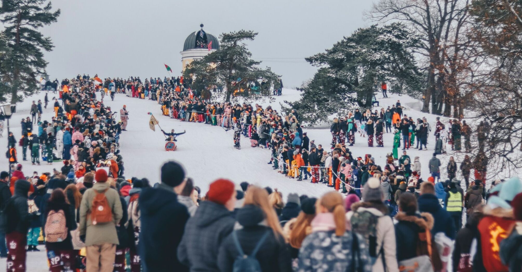 Crowds gather on a snowy hill during a winter festival in Helsinki, surrounded by trees and an observatory in the background