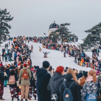 Crowds gather on a snowy hill during a winter festival in Helsinki, surrounded by trees and an observatory in the background