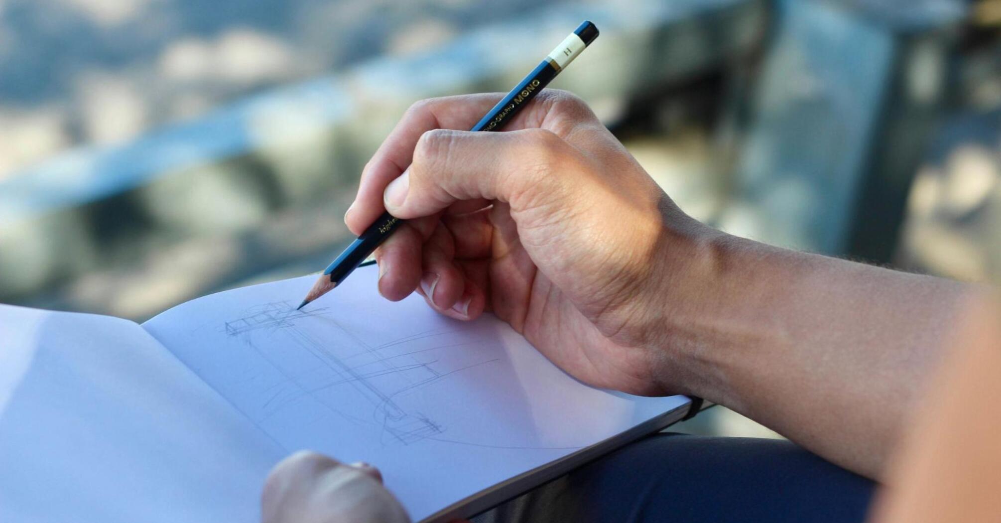 Close-up of a hand sketching in a notebook with a pencil outdoors