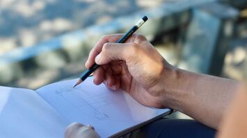 Close-up of a hand sketching in a notebook with a pencil outdoors