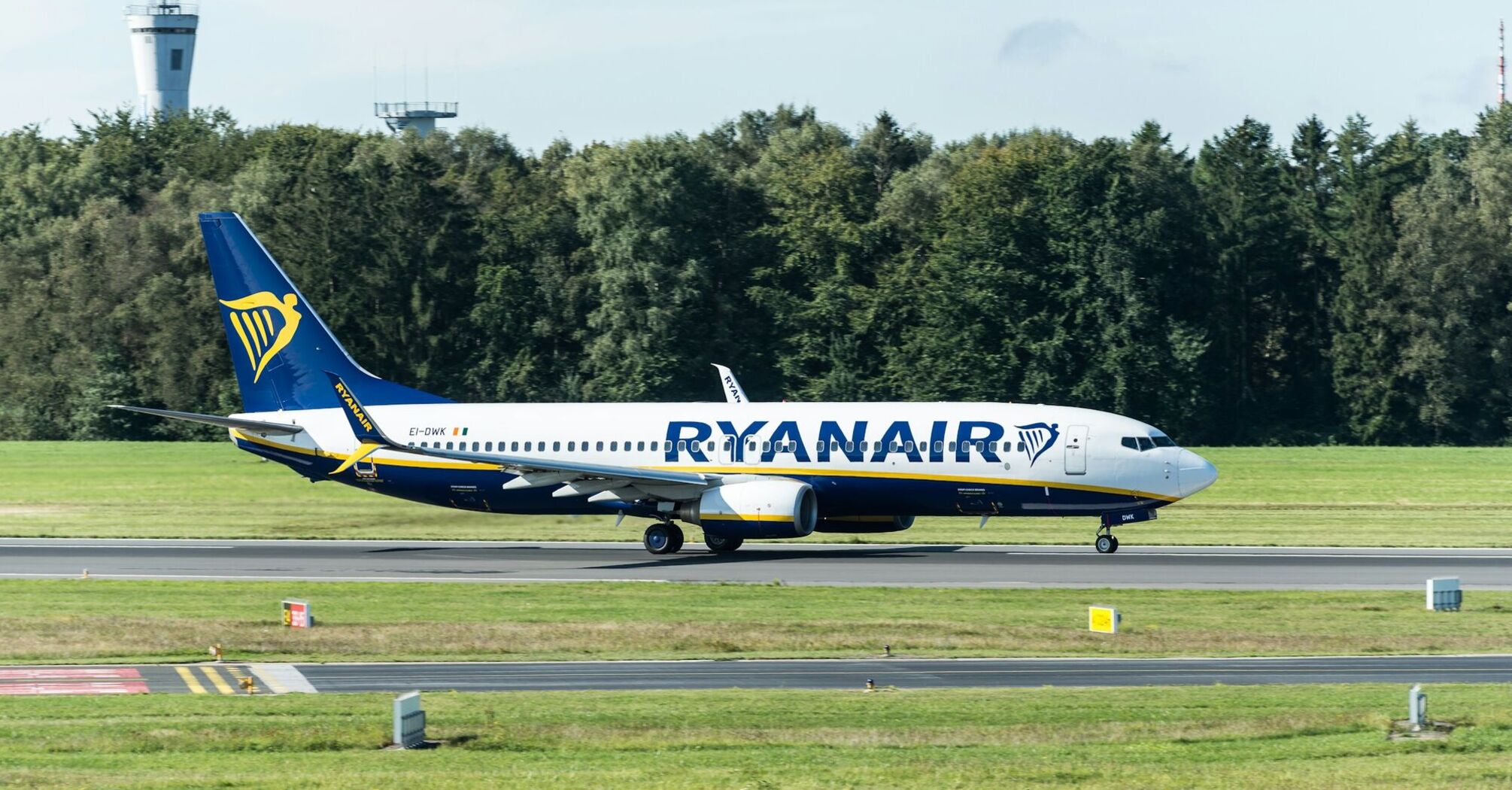 A Ryanair aircraft taxiing on a runway surrounded by greenery and air control towers in the background