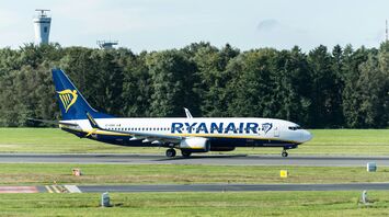 A Ryanair aircraft taxiing on a runway surrounded by greenery and air control towers in the background