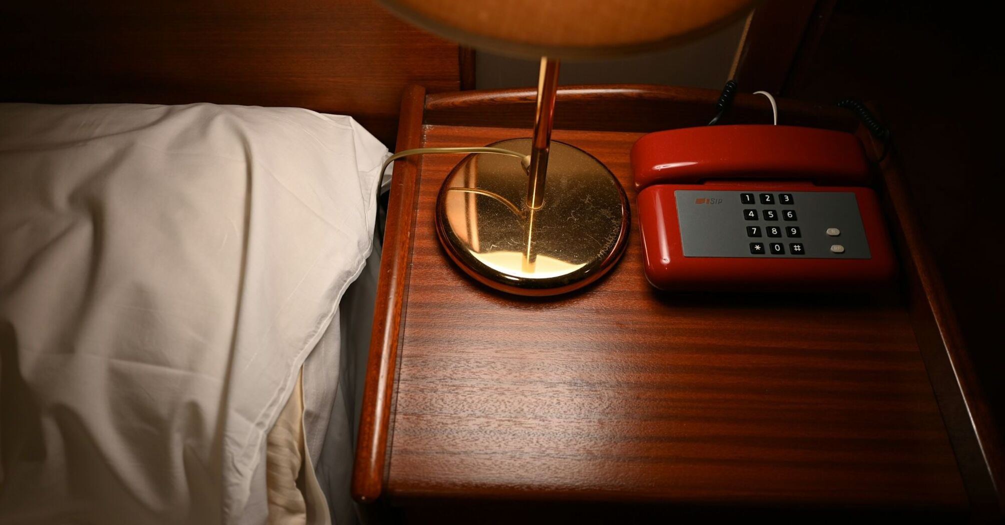 Wooden bedside table with a lamp and red rotary phone beside a white pillow in a dimly lit room