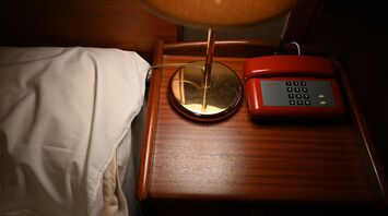 Wooden bedside table with a lamp and red rotary phone beside a white pillow in a dimly lit room