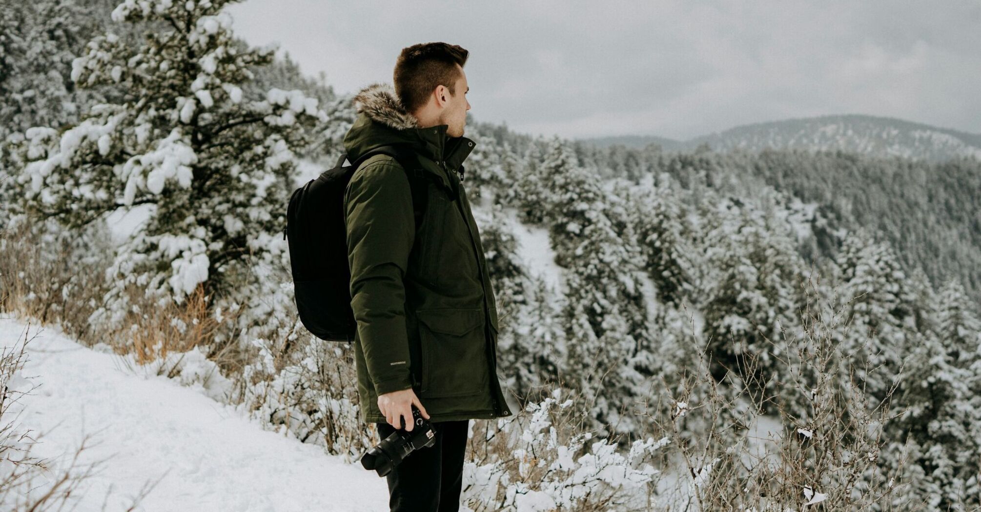 A traveler in a green jacket with a backpack, standing in a snowy forest landscape, holding a camera, gazing at the scenic winter view