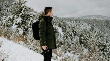 A traveler in a green jacket with a backpack, standing in a snowy forest landscape, holding a camera, gazing at the scenic winter view