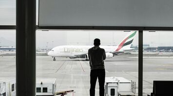 A person looking at an Emirates airplane through a glass window at the airport