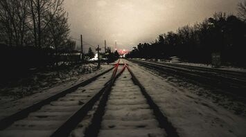 Snow-covered railway tracks illuminated by red signal lights under a cloudy evening sky