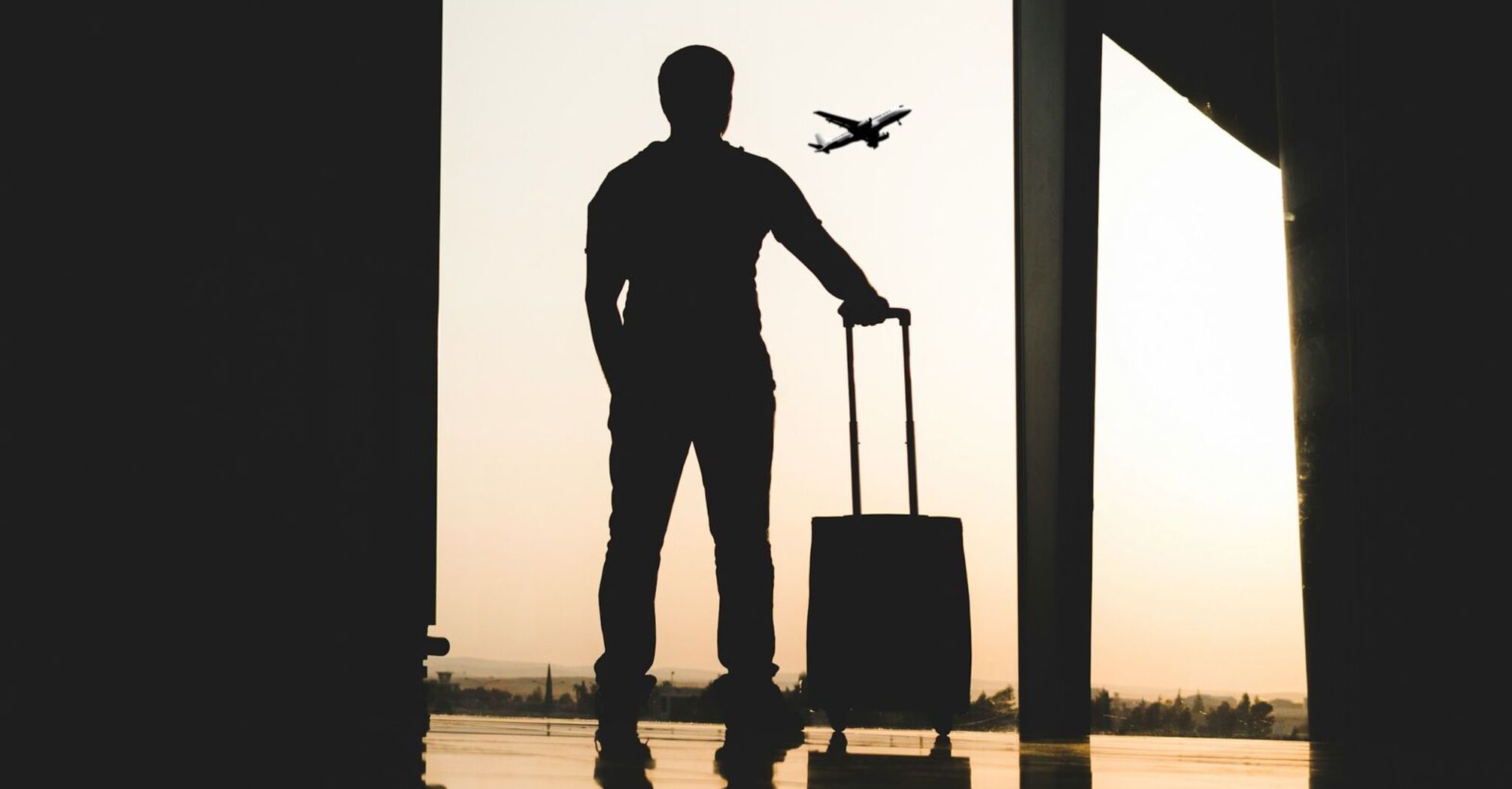 A silhouette of a traveler with a suitcase standing by a window at an airport, with an airplane flying in the background