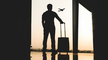 A silhouette of a traveler with a suitcase standing by a window at an airport, with an airplane flying in the background