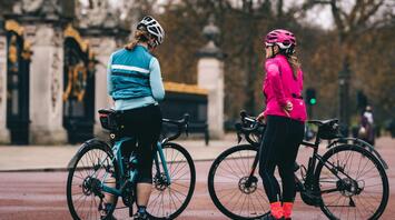 Two cyclists in bright safety gear stopped on a road near a gate, discussing something