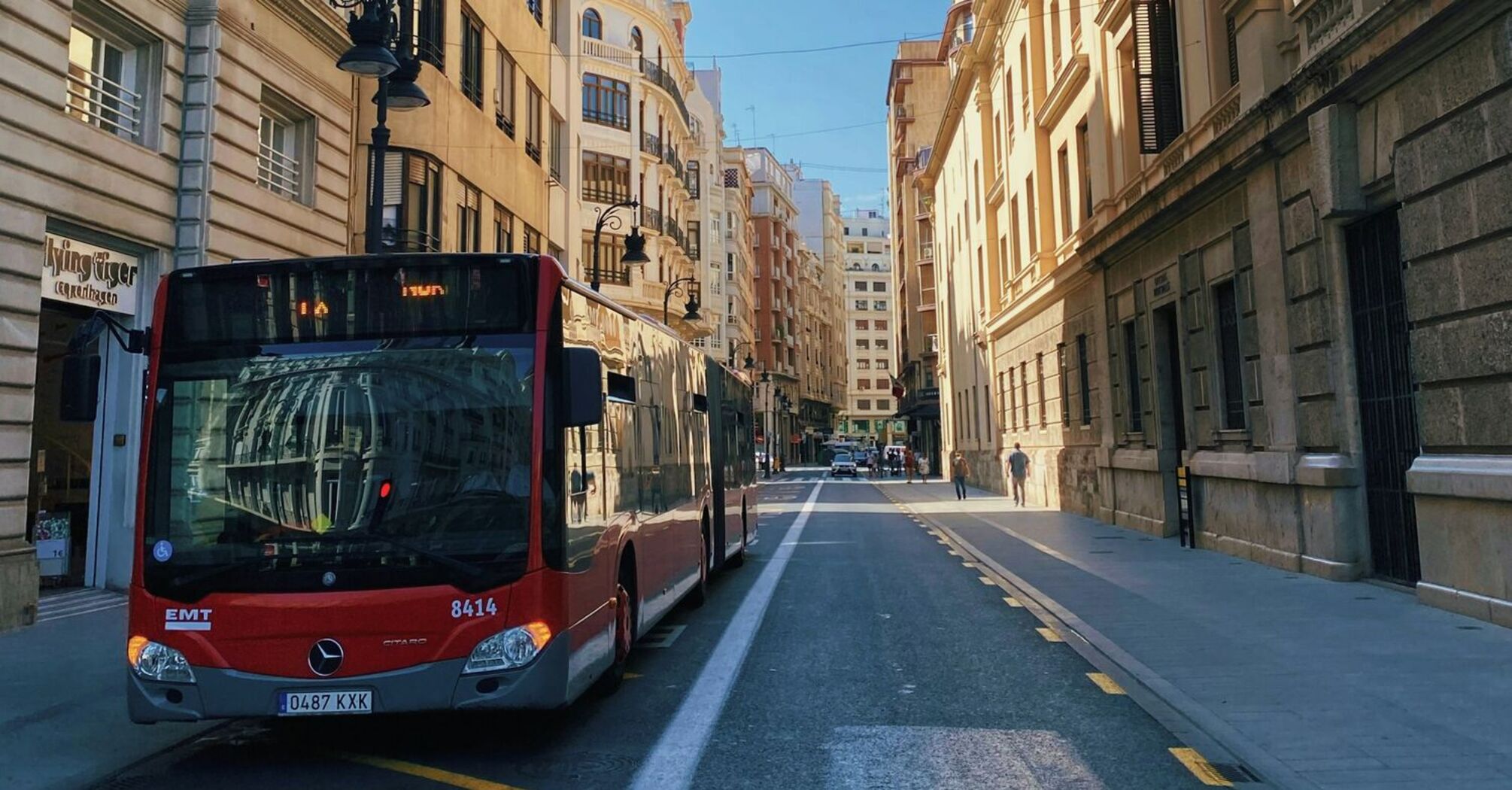 A red city bus on a quiet urban street in Spain surrounded by historic buildings under a clear blue sky