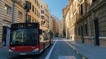 A red city bus on a quiet urban street in Spain surrounded by historic buildings under a clear blue sky