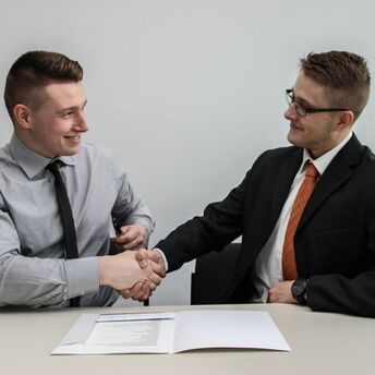 Two men in formal attire shaking hands over a signed agreement at a table
