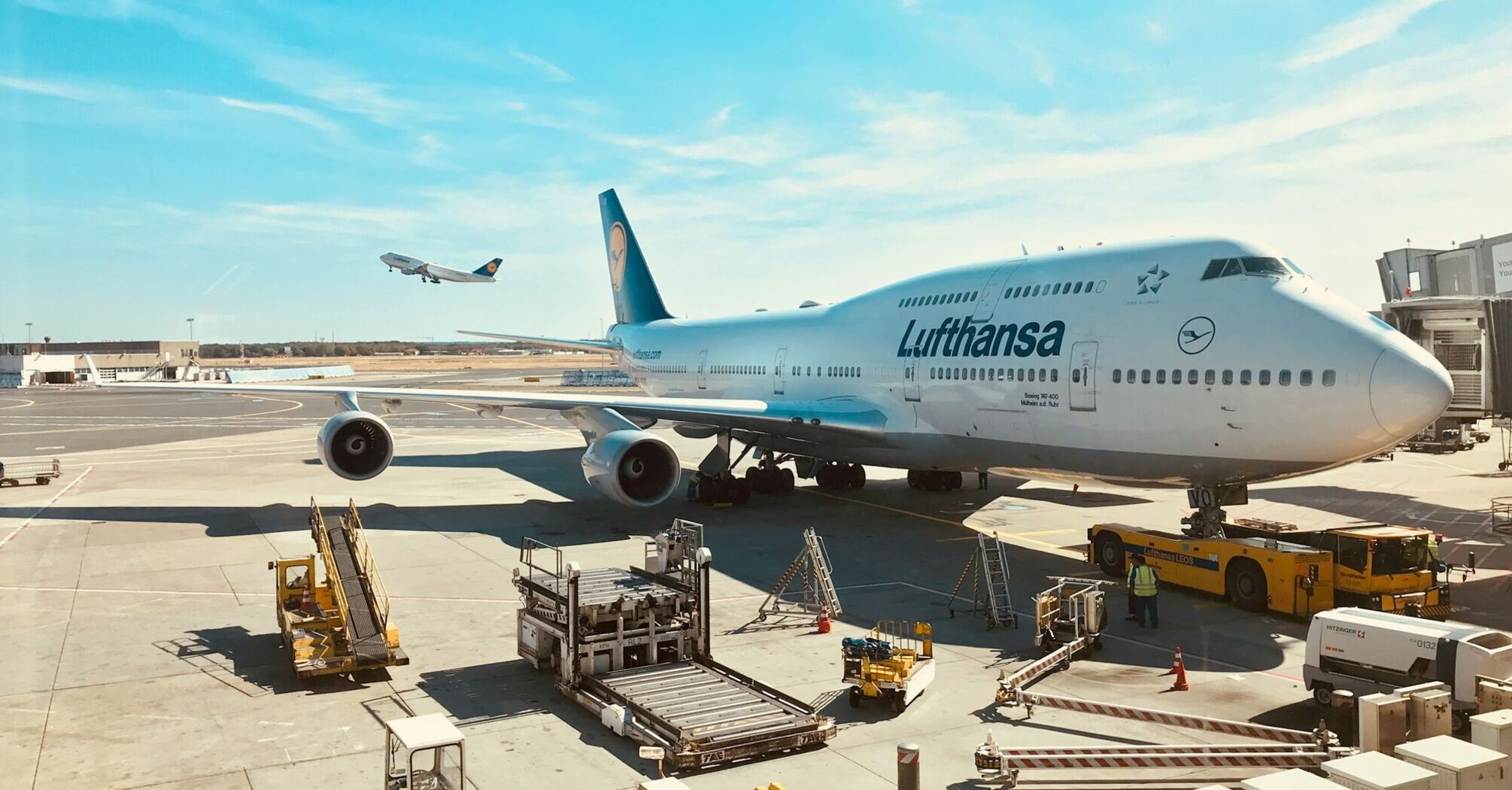 Lufthansa aircraft parked at an airport with ground crew and a plane taking off in the background