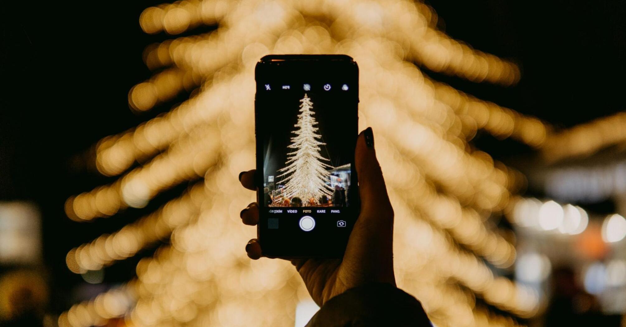 A smartphone captures a brightly lit Christmas tree surrounded by blurred festive lights