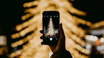 A smartphone captures a brightly lit Christmas tree surrounded by blurred festive lights