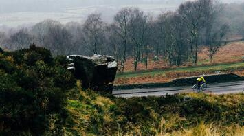 A cyclist rides along a quiet rural road surrounded by misty trees and rocky hills