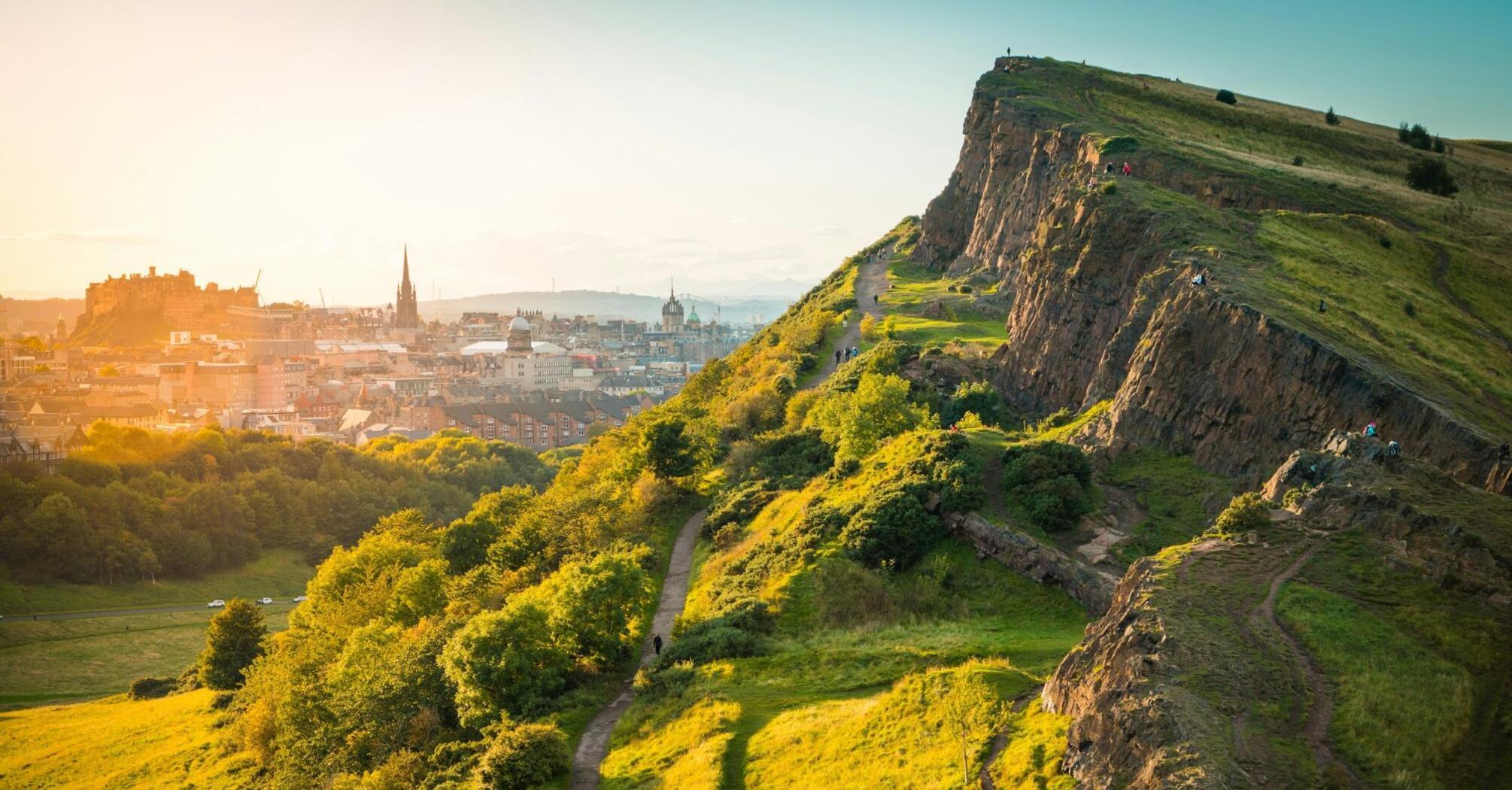 View of Edinburgh cityscape from Arthur's Seat, featuring a green hill and historic skyline at sunset