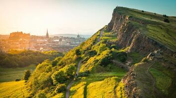 View of Edinburgh cityscape from Arthur's Seat, featuring a green hill and historic skyline at sunset