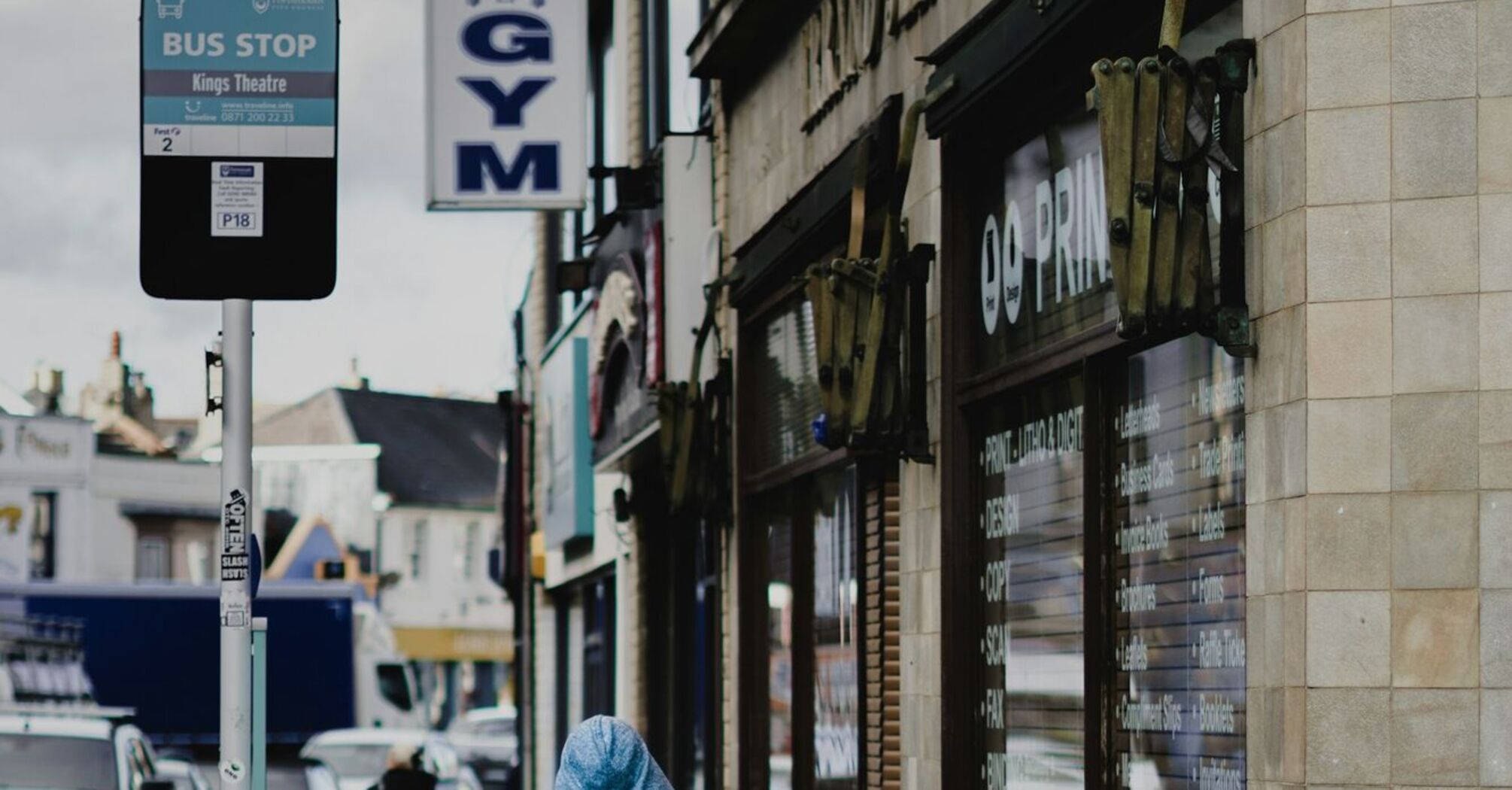 A person in a blue cloak walking near a bus stop labeled "Kings Theatre" in Portsmouth, with nearby shops visible