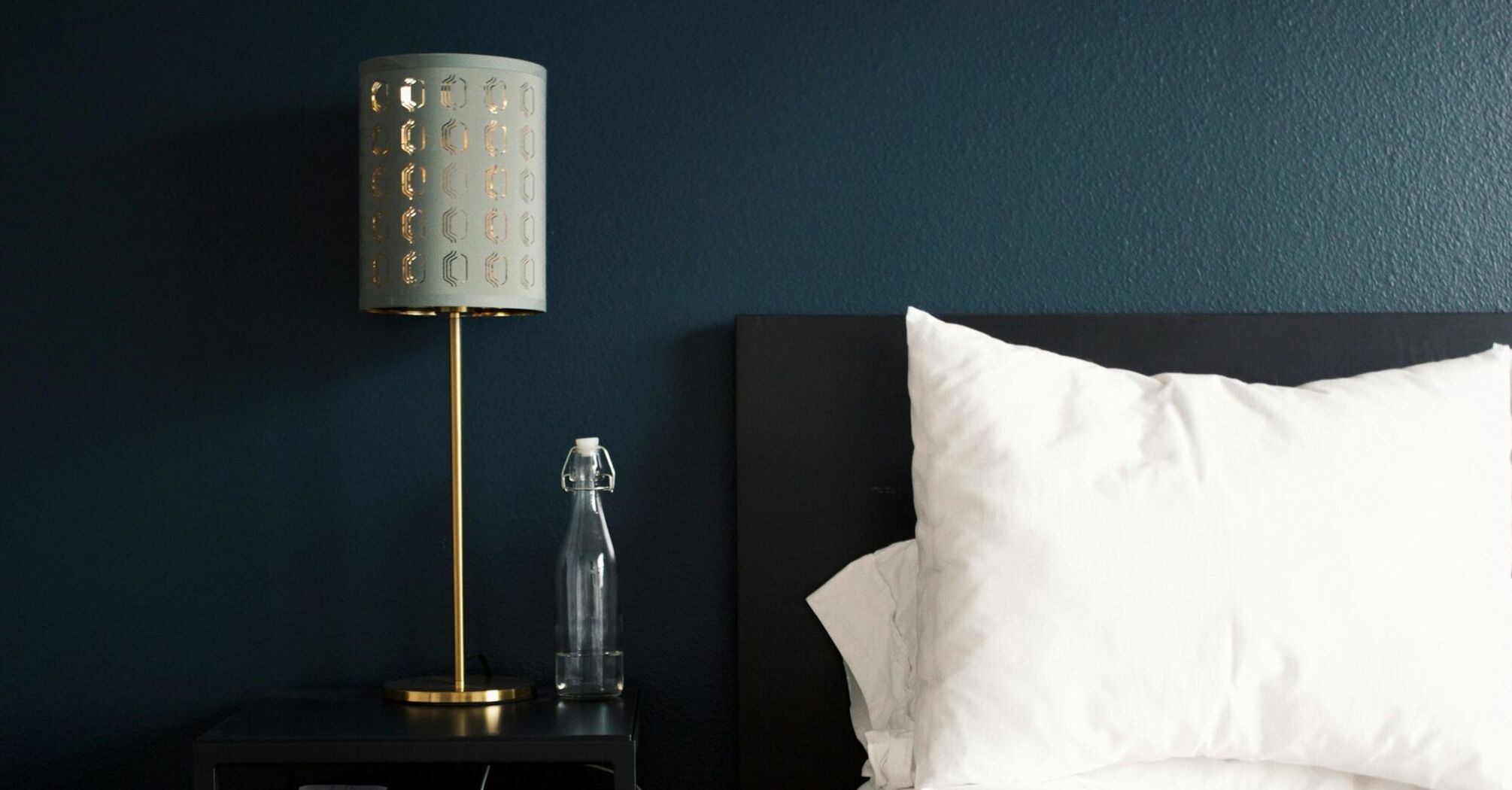 A neatly arranged hotel bedside table with a modern lamp, a glass water bottle, and a white pillow on a bed