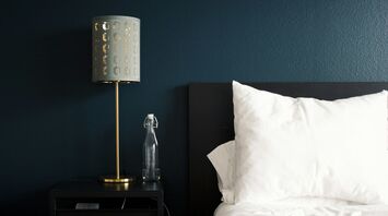 A neatly arranged hotel bedside table with a modern lamp, a glass water bottle, and a white pillow on a bed