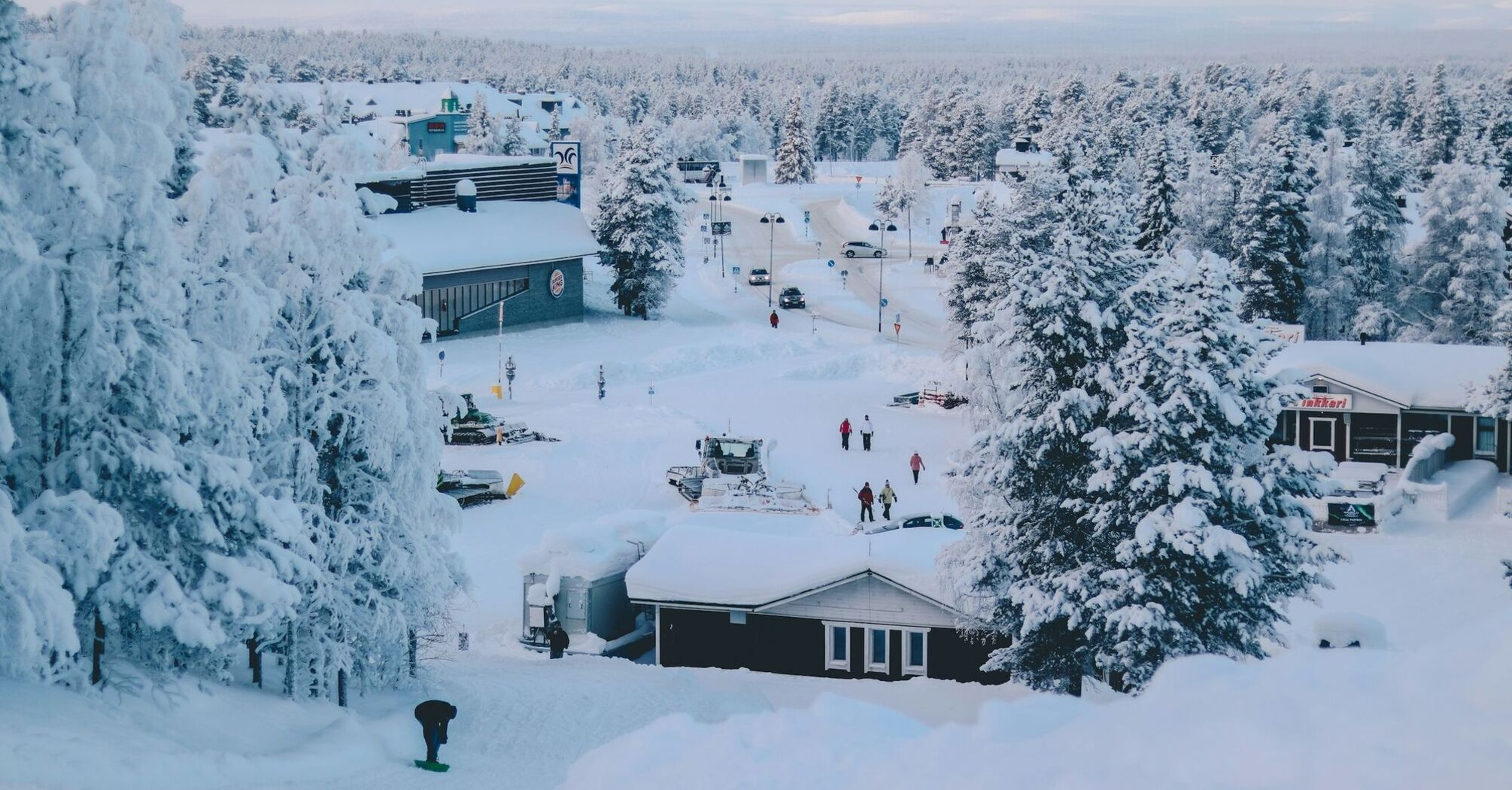 Snow-covered village in Finnish Lapland with trees, small buildings, and people walking through the winter landscape