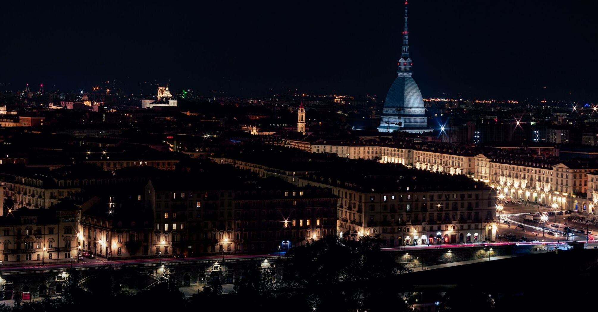 A nighttime view of Turin, Italy, featuring the illuminated Mole Antonelliana and surrounding cityscape