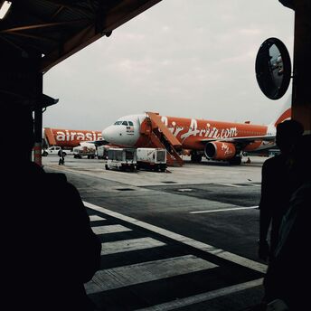 An AirAsia aircraft parked at the terminal under cloudy skies, with visible ground equipment and a silhouette of passengers in the foreground