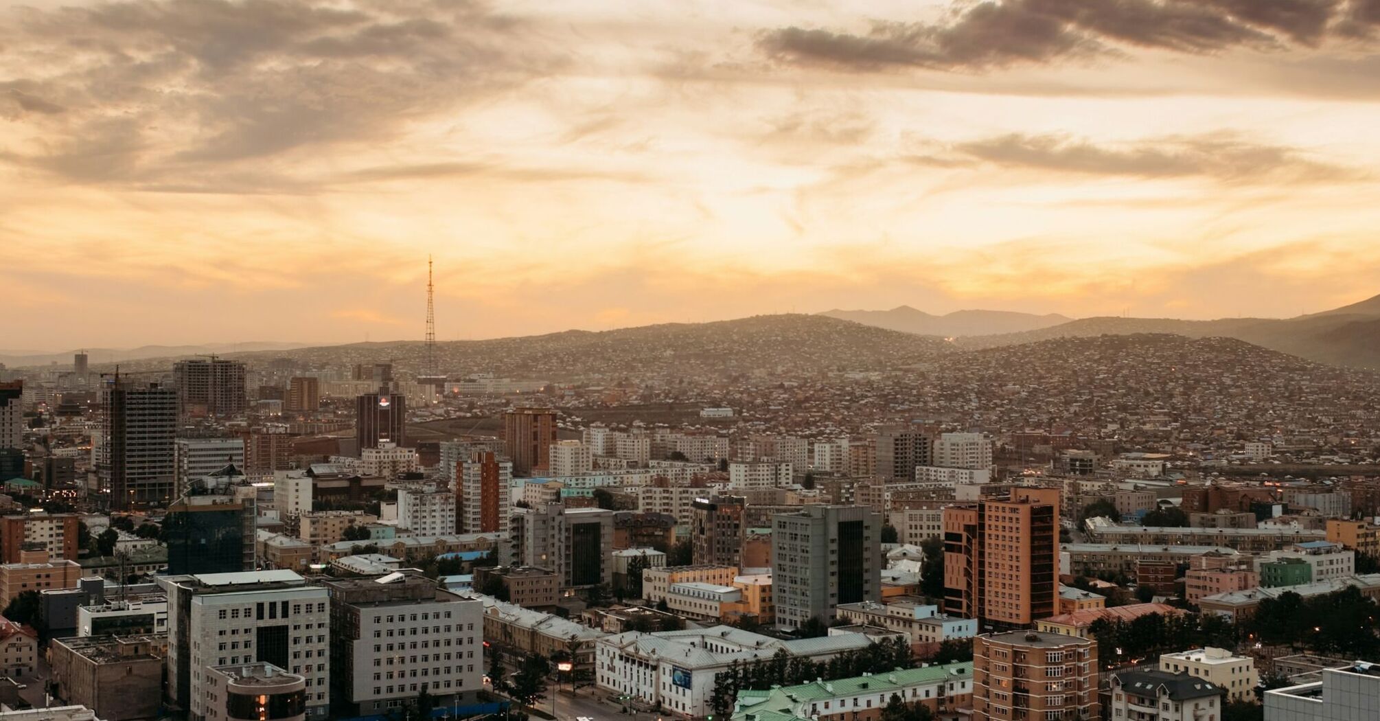 Aerial view of Ulaanbaatar, Mongolia's capital, with a blend of modern buildings and surrounding hills under a golden sunset sky