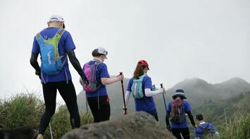 A group of hikers walking along a mountain trail, wearing backpacks and using trekking poles, with misty mountains in the background