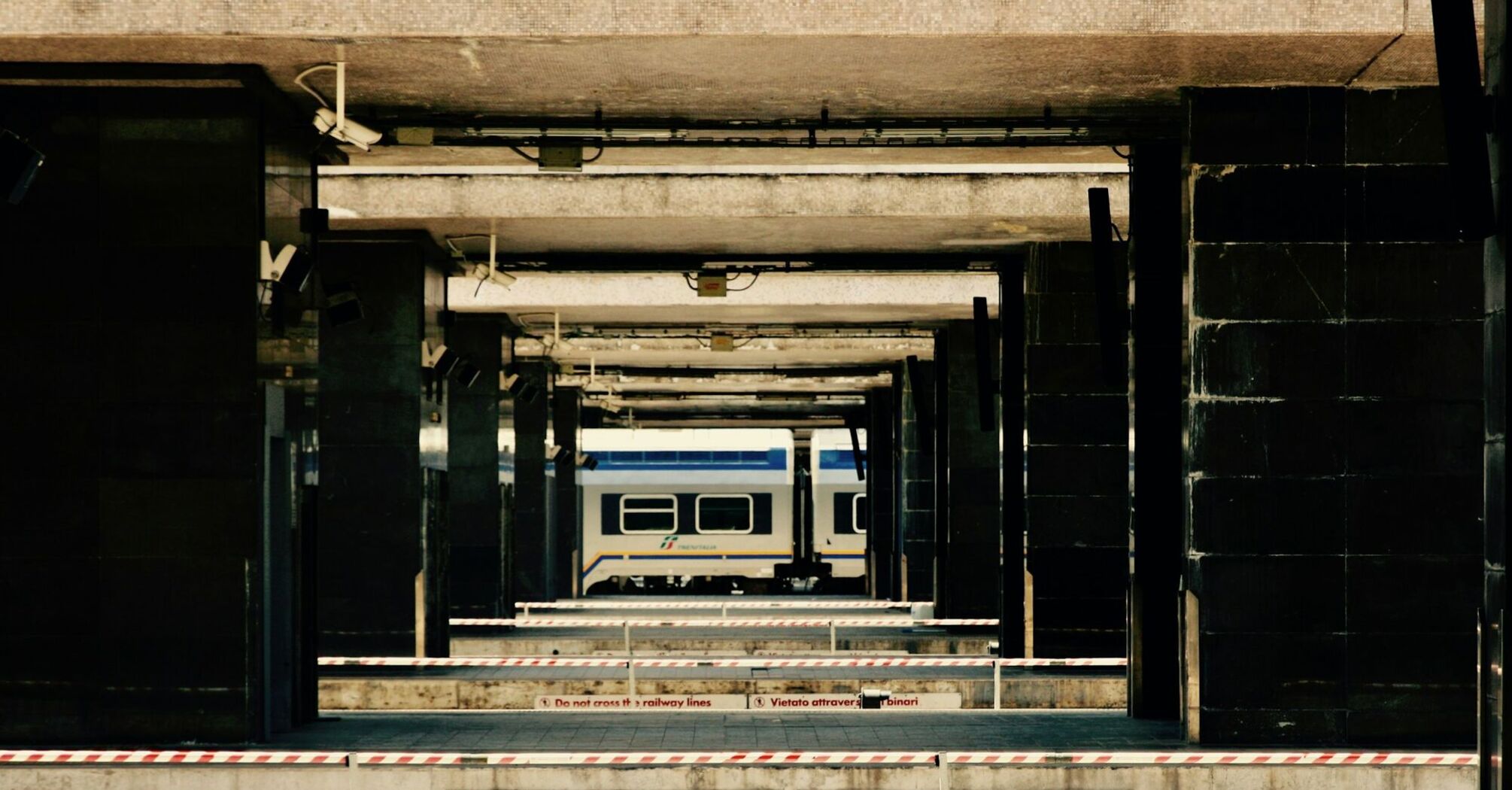 A train platform in Italy with a stationary Sicilia Express train visible in the background and barriers marked with safety warnings in Italian and English