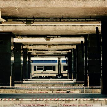 A train platform in Italy with a stationary Sicilia Express train visible in the background and barriers marked with safety warnings in Italian and English